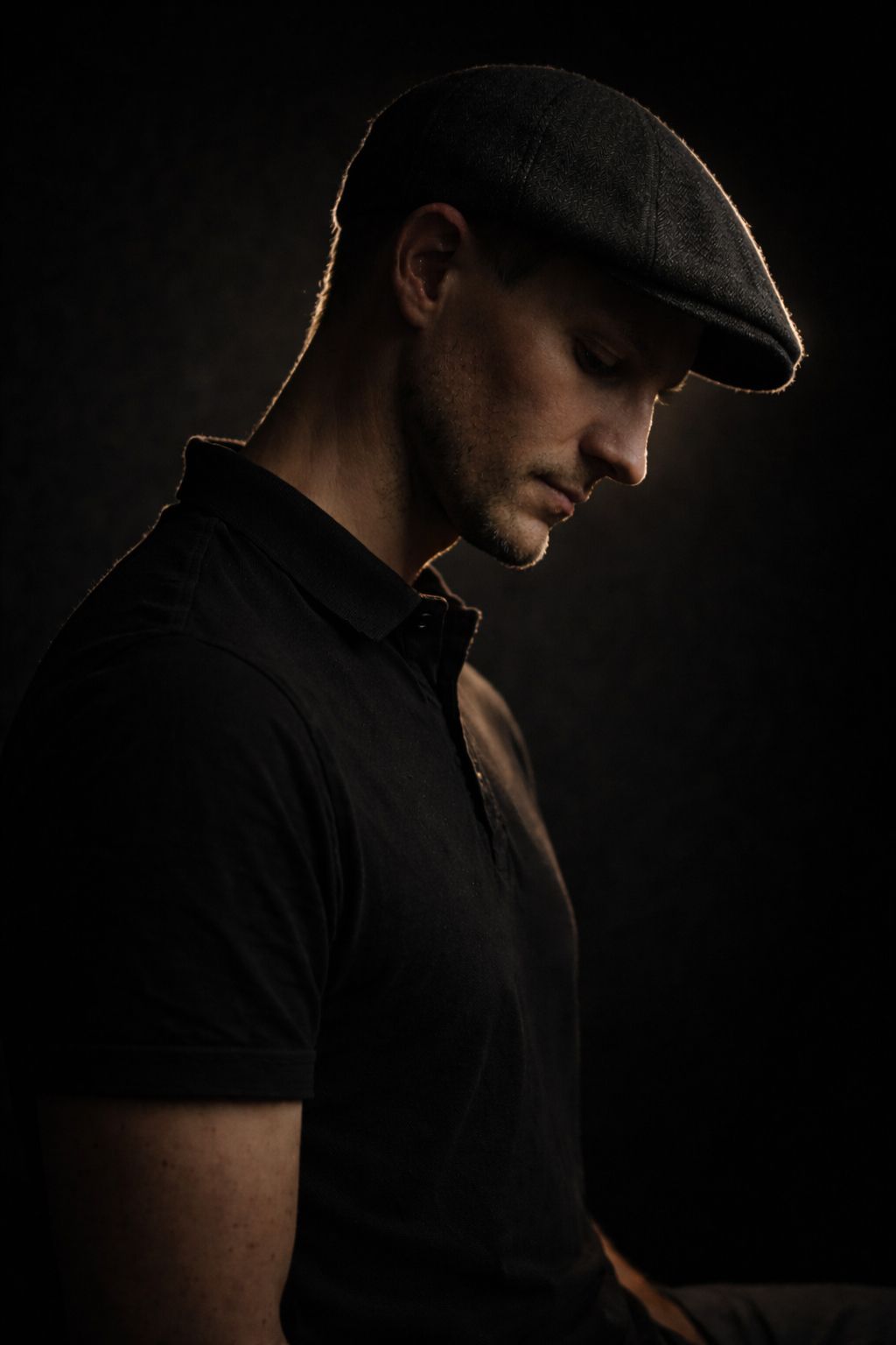 A brooding man in a dark flat cap and black polo shirt gazes downward against a dramatic dark background.