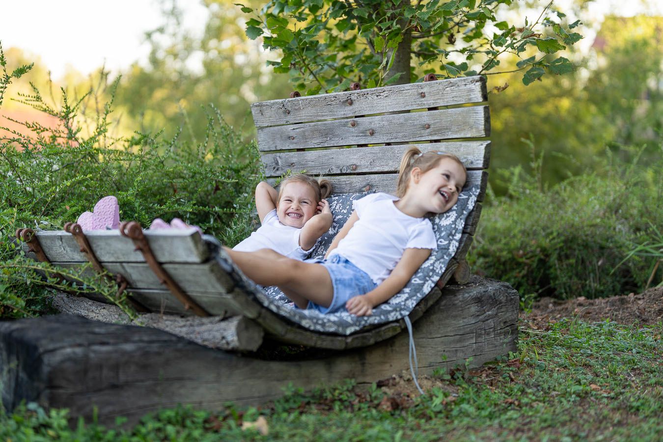 Two young girls relaxing and laughing together in a rustic wooden outdoor hammock chair surrounded by greenery.