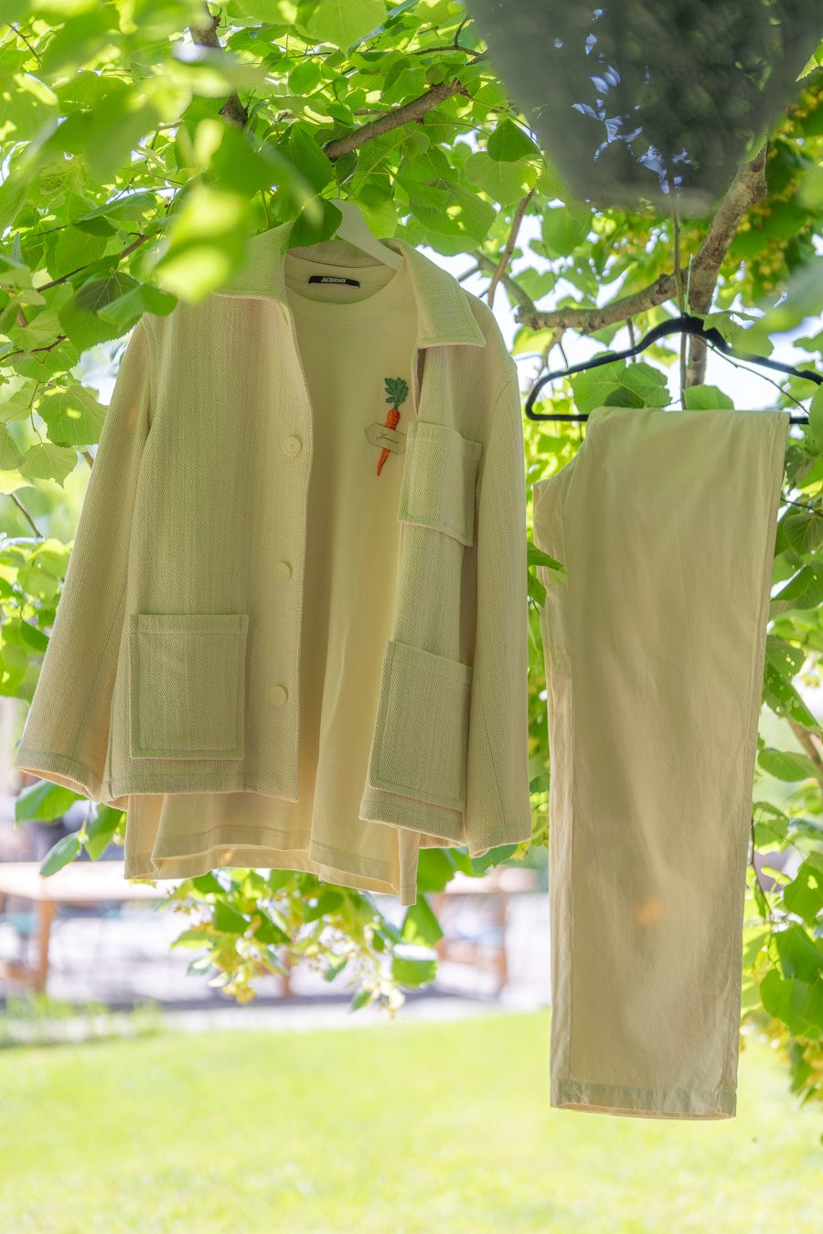 Yellow linen jacket and pants set hanging on a hanger outdoors among green tree branches in sunlight.