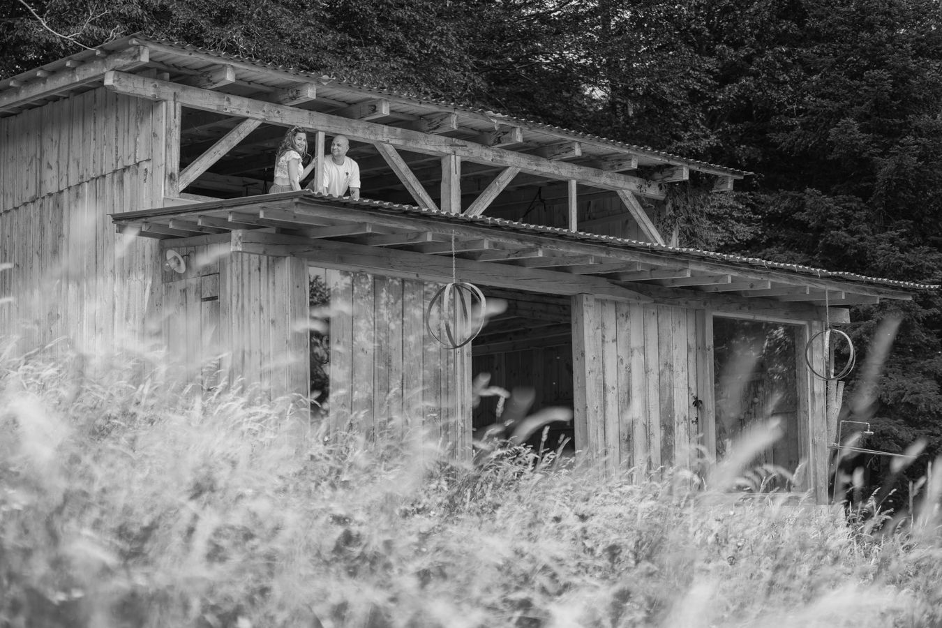 Two people stand on the upper level of a rustic wooden barn surrounded by tall grass, captured in black and white.
