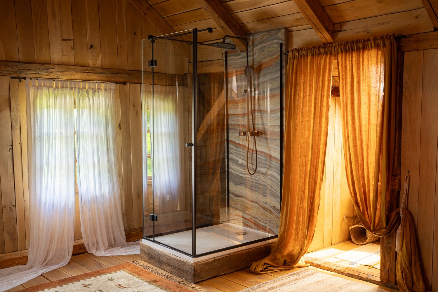 Modern rustic bathroom with glass shower enclosure, wooden walls, and golden curtains creating a warm atmosphere.