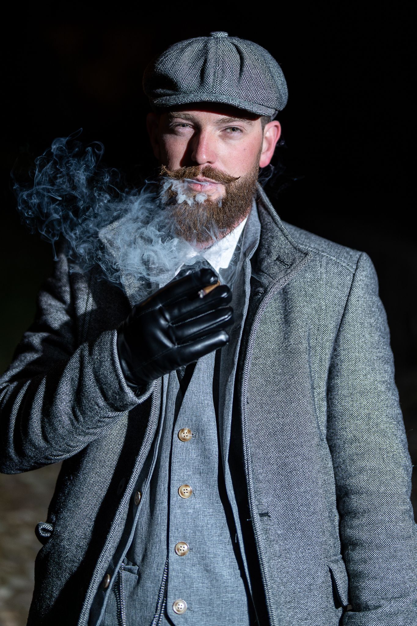 Person in vintage grey coat and cap exhaling smoke against dark background.
