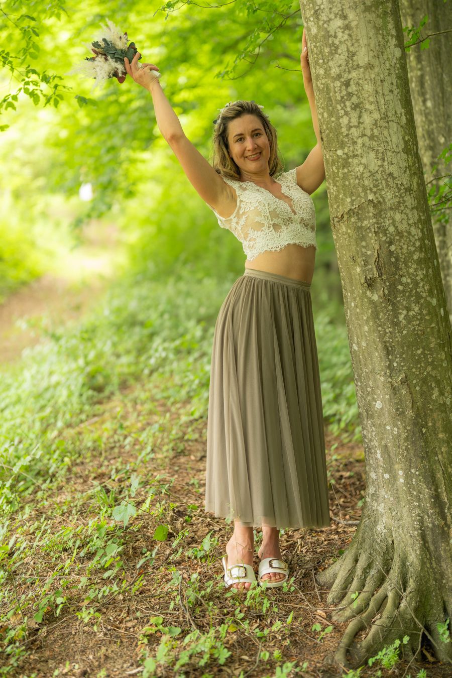 Woman in lace top and maxi skirt holding bouquet aloft beside a tree in a lush green forest path.