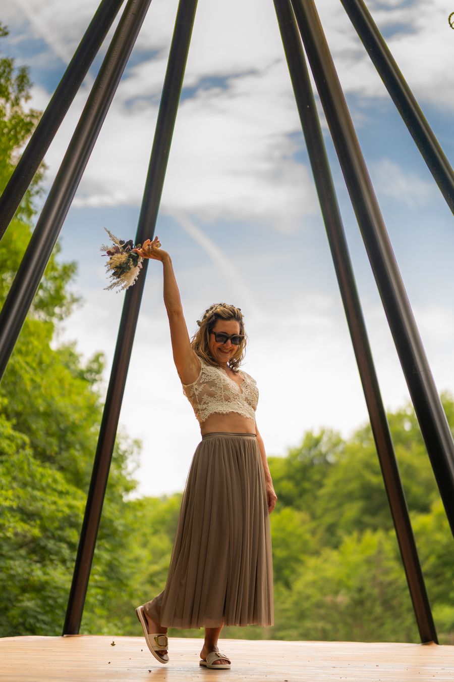 Bride in lace top and tulle skirt raising bouquet under teepee frame with green trees and blue sky.