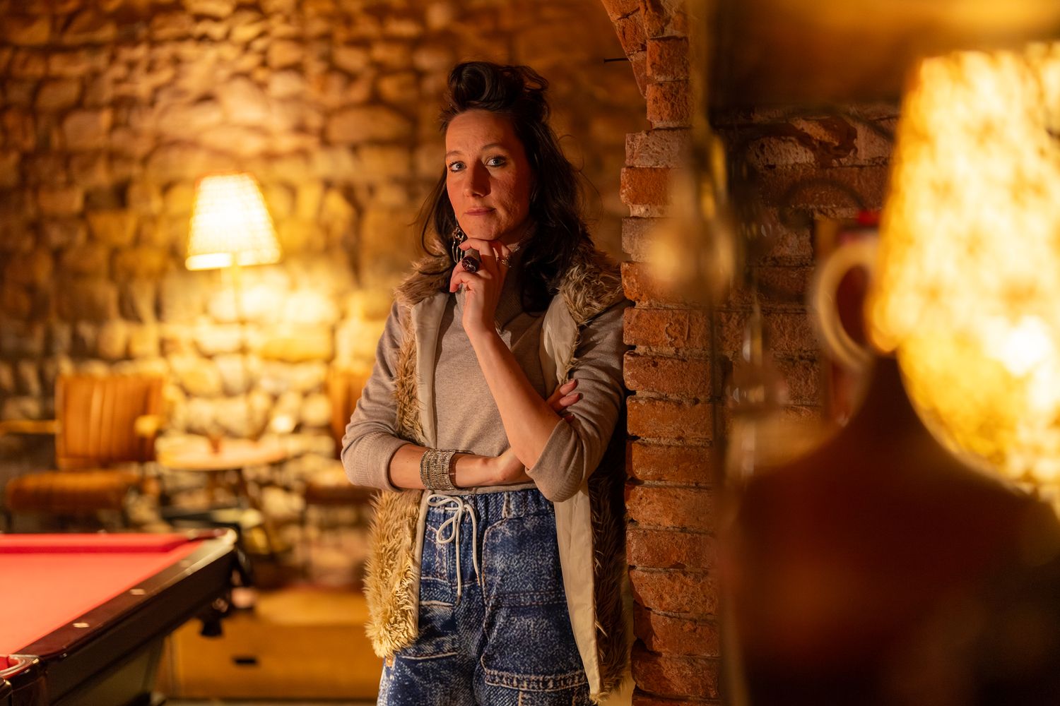 Person in denim overalls poses in atmospheric brick cellar with warm lighting and vintage decor.