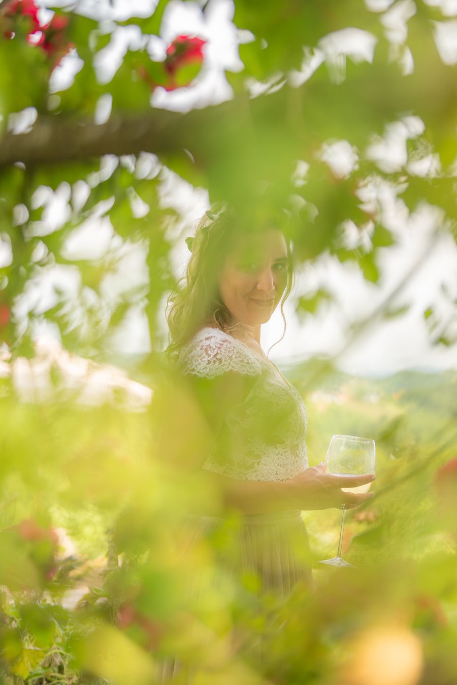 Woman in white dress holding a wine glass surrounded by lush green foliage and red flowers in a garden.