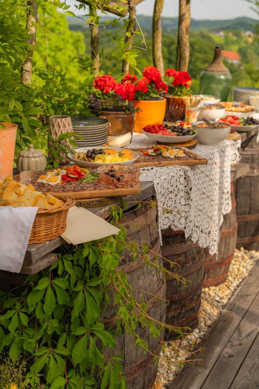 Rustic outdoor buffet table on wine barrels with assorted foods, red flowers, and lace cloth amid lush greenery.
