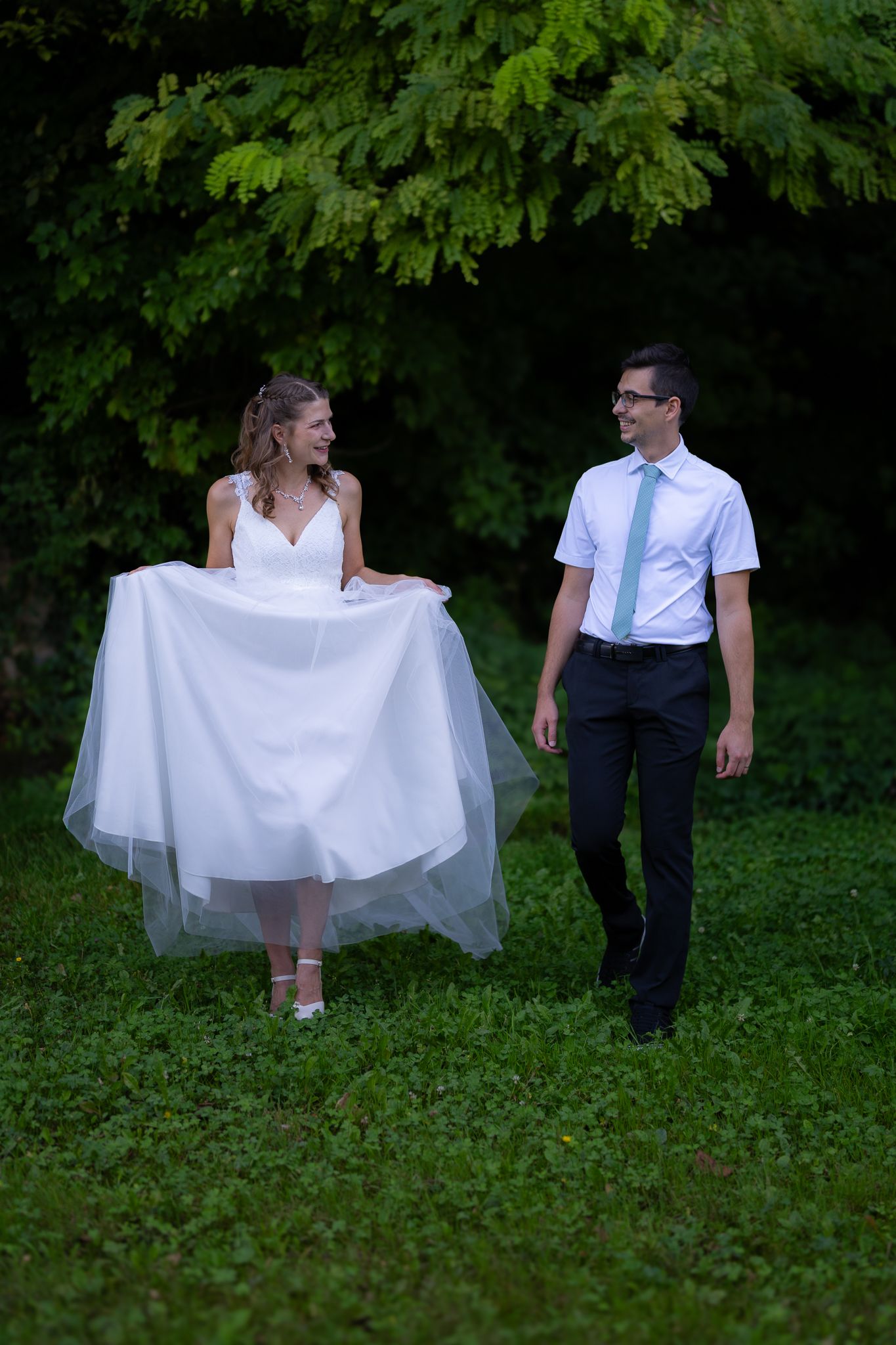 A bride in a white gown and groom walk together on grass surrounded by trees.
