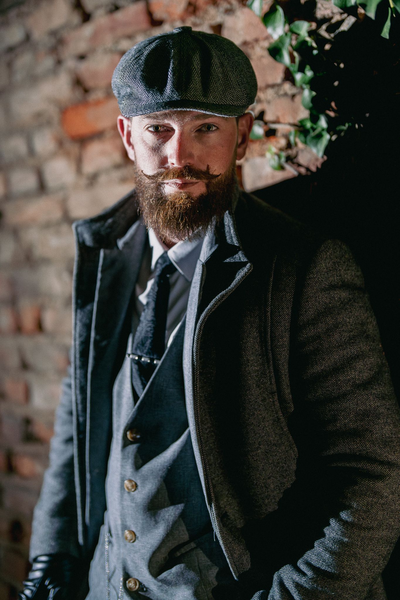 Man in vintage-style clothing with flat cap and beard poses against brick wall in moody lighting.