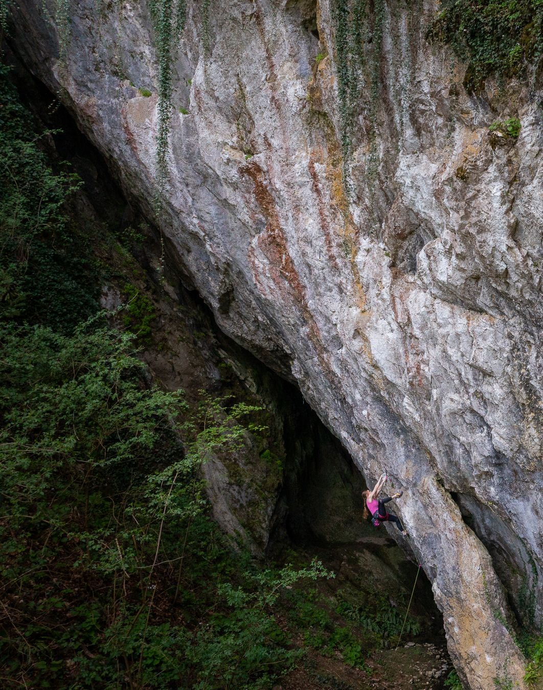 Rock climber scaling an overhanging limestone cave formation in a forested setting.