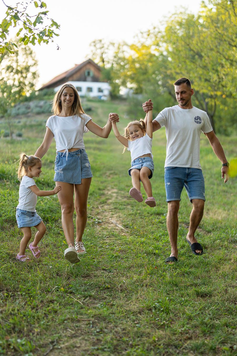 Happy family in matching white tees and denim walking outdoors, parents swinging young child while another child holds hands.