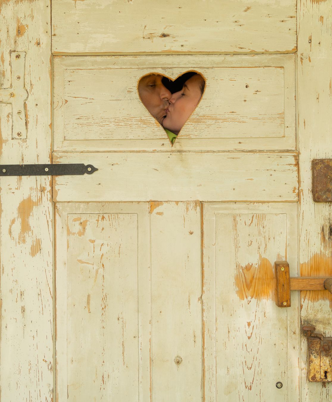 A couple kissing through a heart-shaped cutout in a rustic, weathered white wooden door.