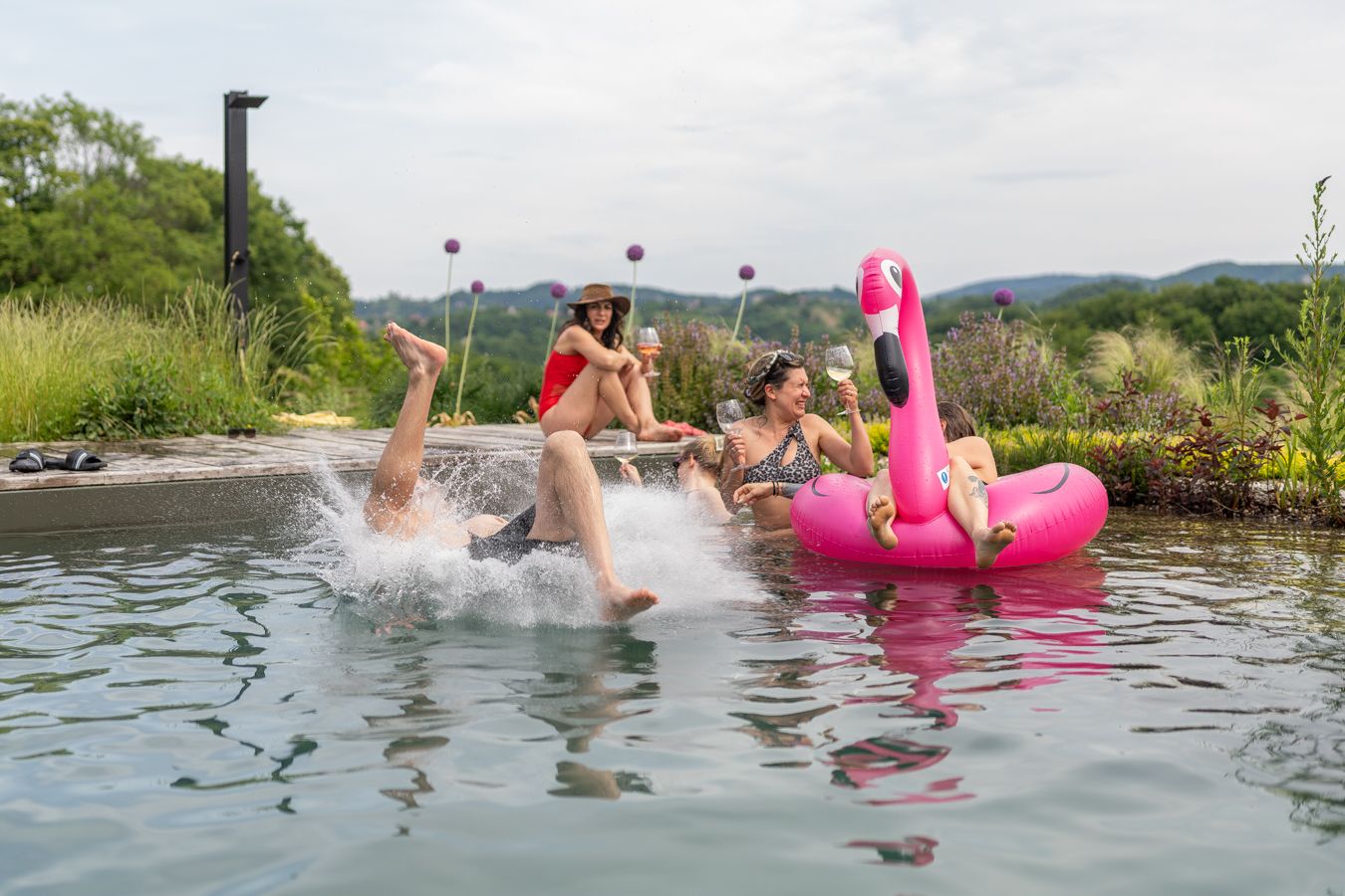 People enjoying a pool party with a pink flamingo float and water splashing on a sunny day.