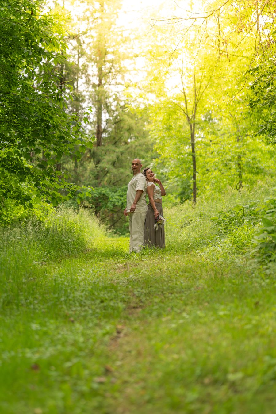 Couple standing together on a lush green forest path, bathed in warm golden sunlight filtering through the trees.