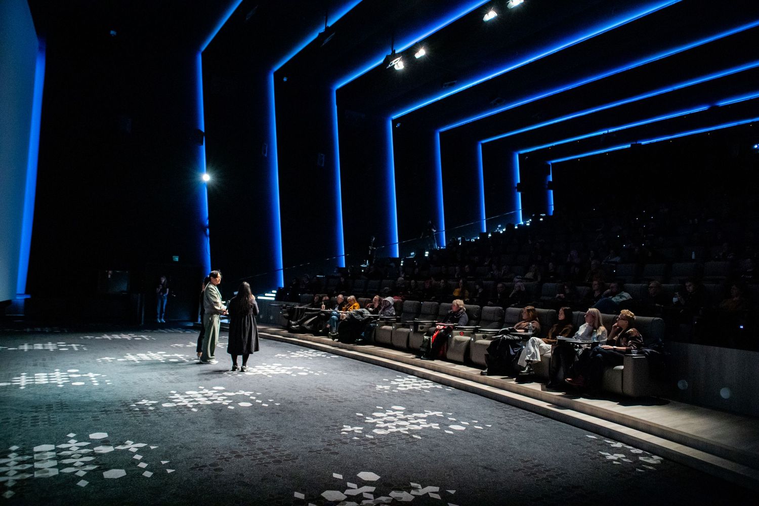 Speaker addressing a seated audience in a modern blue-lit cinema auditorium at a film event.