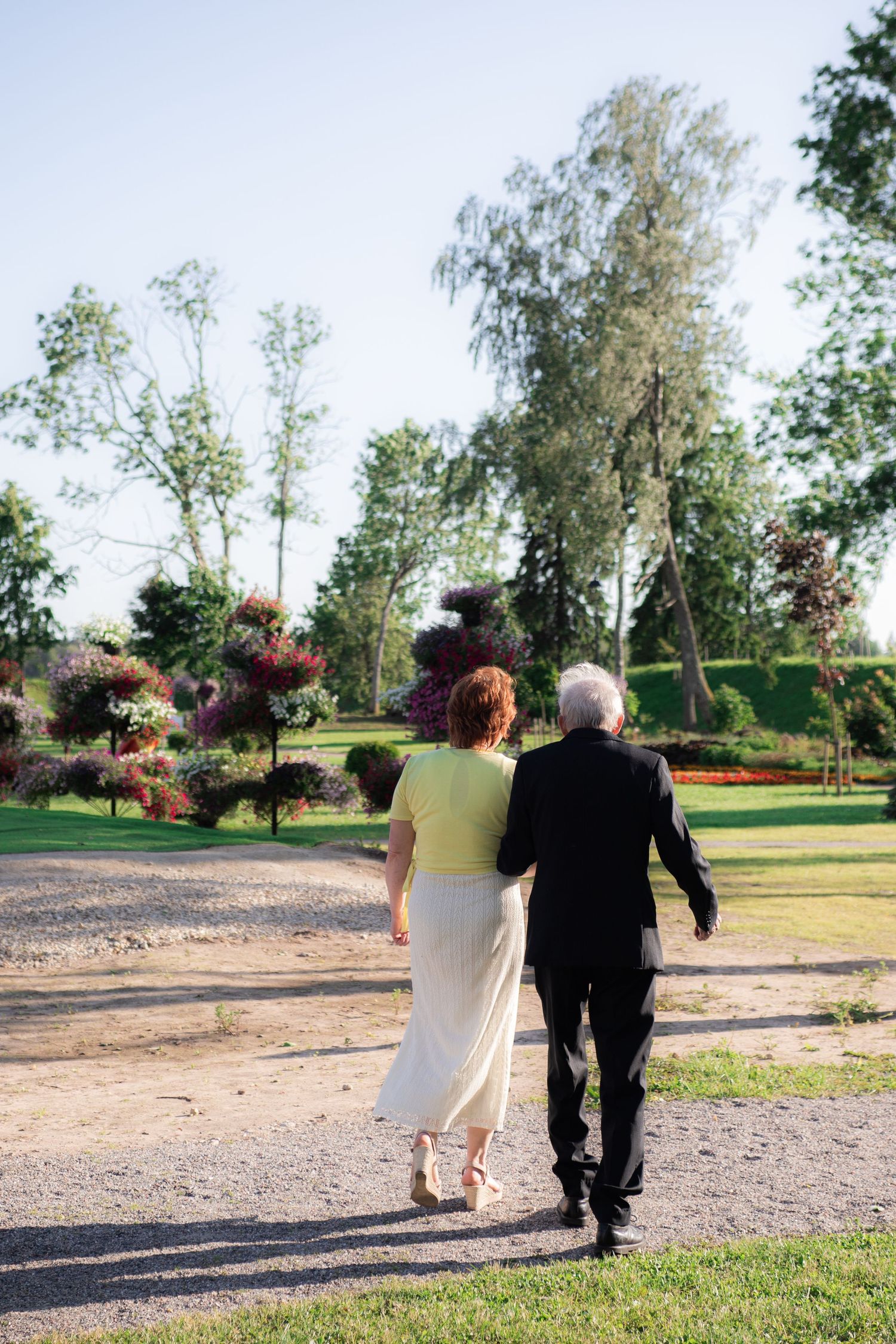 Elderly man and woman walking arm in arm through a scenic garden path on a sunny day.