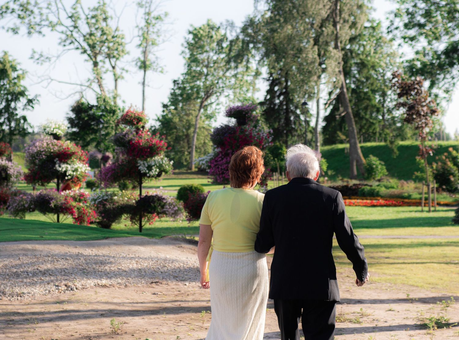 Elderly couple walking together through a lush green garden with colorful flowering trees.