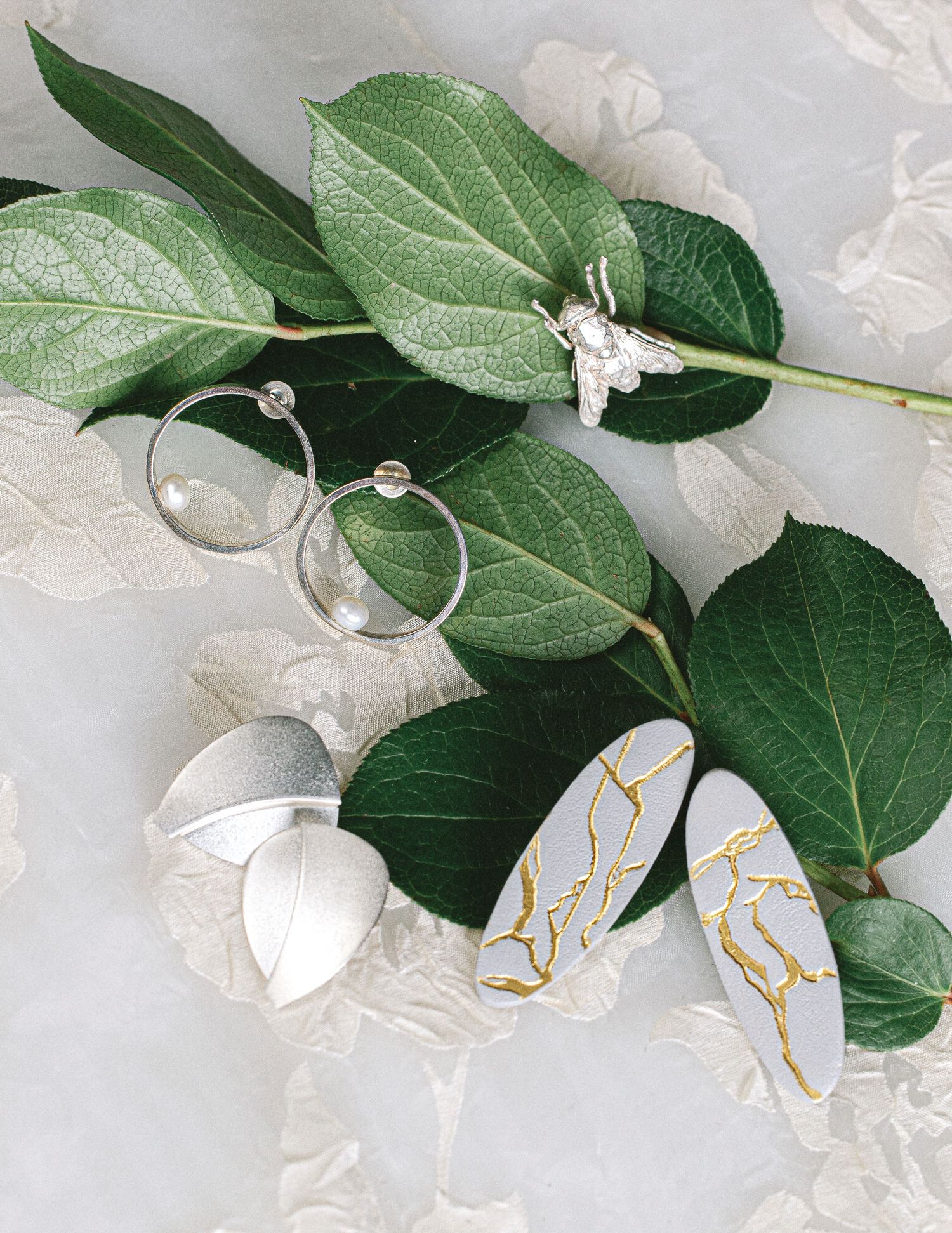 Silver hoop earrings, butterfly ring, and gold-veined gray oval earrings arranged on green eucalyptus leaves.