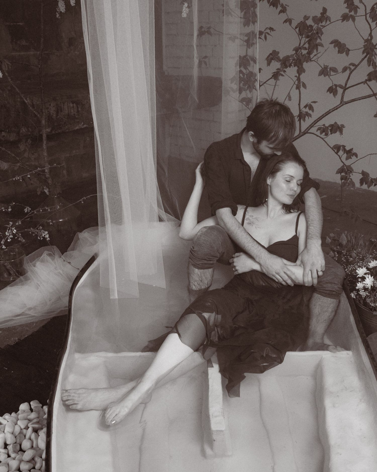 Black and white photo of a couple sitting intimately together in a bathtub, surrounded by soft drapery and plants.