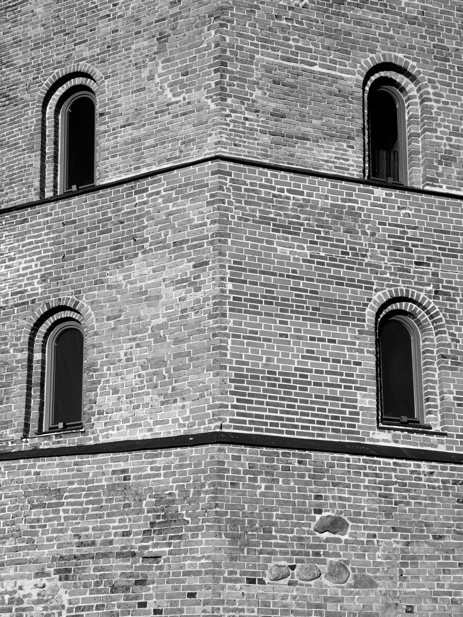 Black and white close-up of a brick tower corner featuring four arched windows with detailed brickwork.