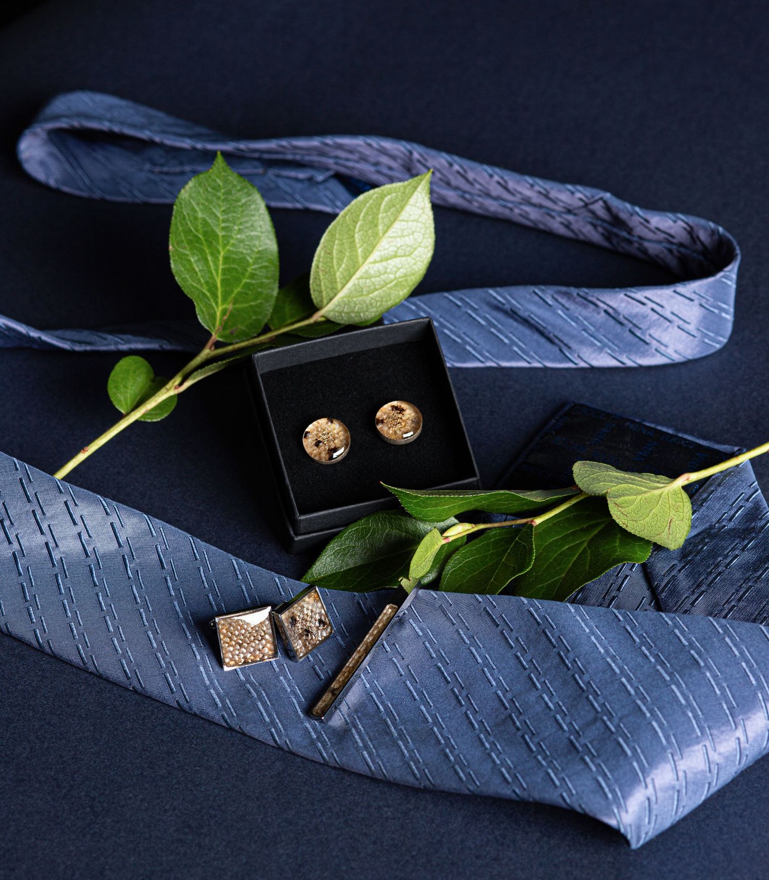 Gold cufflinks in a black box styled with a blue striped tie and green leaves.
