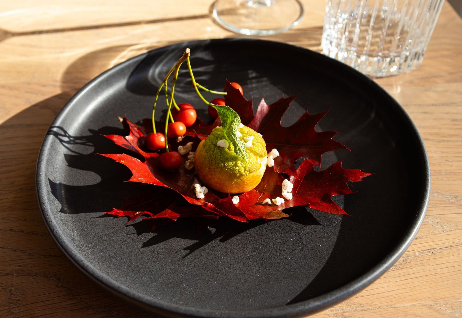 Elegant fall-inspired plating featuring vibrant red maple leaves, citrus fruit, herb sorbet, and scattered berries on black plate.