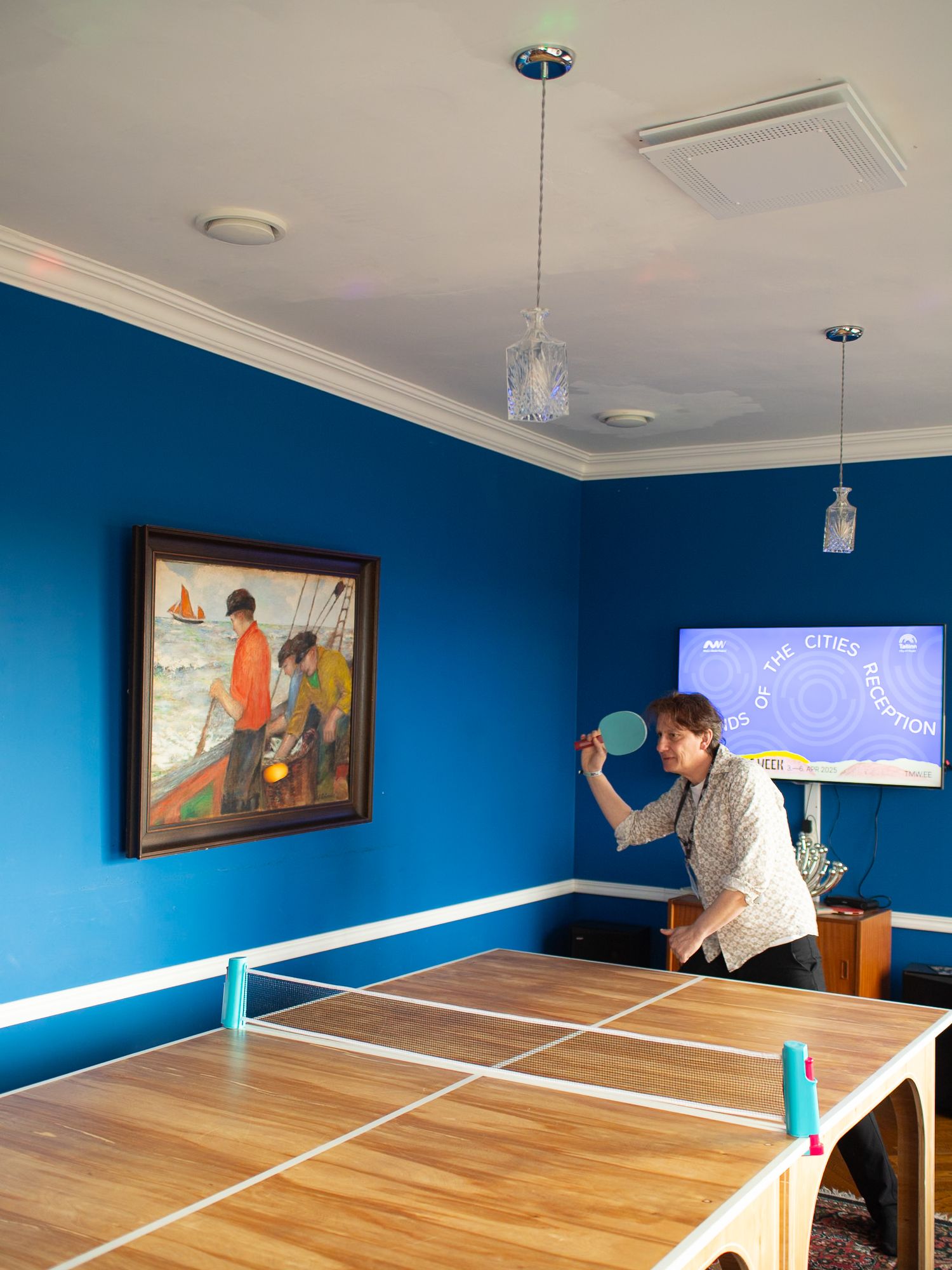 Woman playing ping pong in a blue room with a painting on the wall and a large screen displaying text.