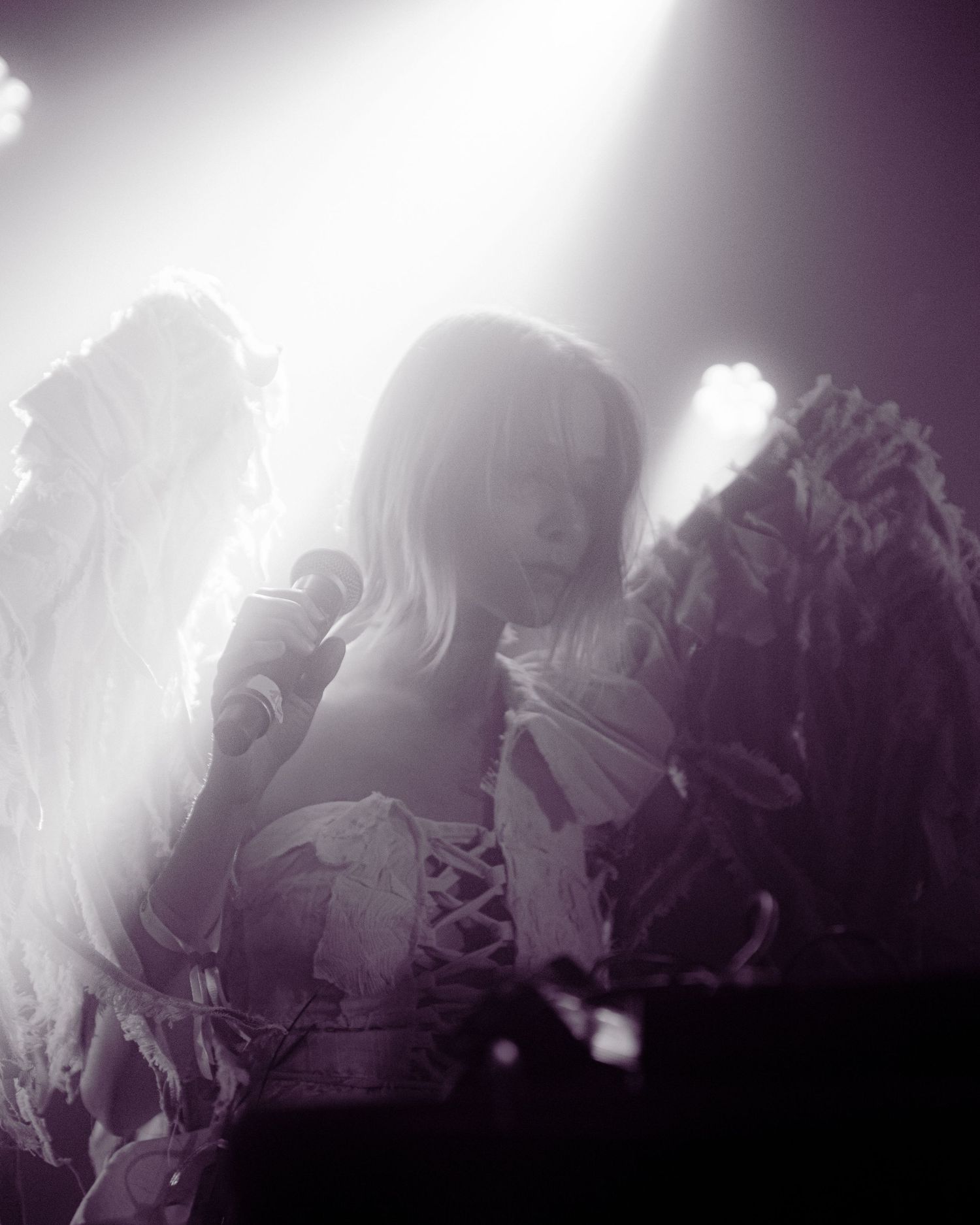 Black and white photo of a blonde performer with angel wings singing into a microphone under bright stage lights.