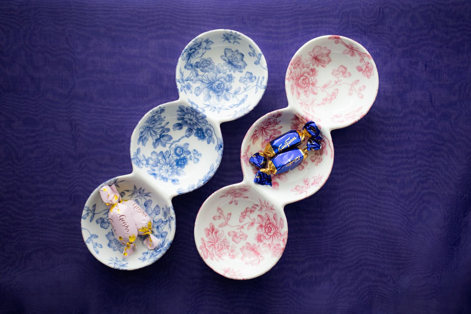 Six floral ceramic bowls in blue and pink patterns displayed on purple fabric, containing wrapped candies and a small figurine.