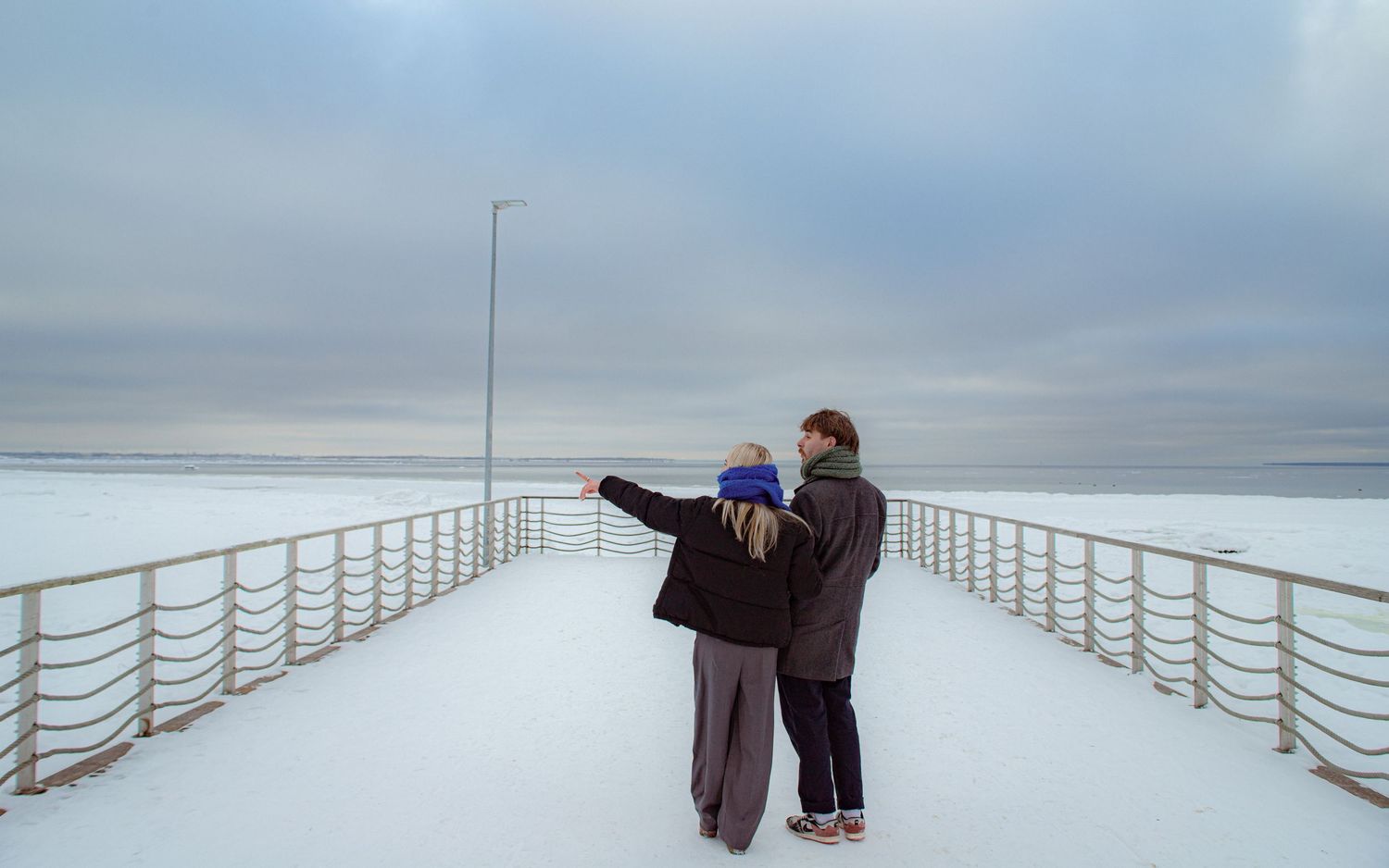 Couple standing on a snow-covered bridge overlooking a frozen winter landscape under a cloudy sky.
