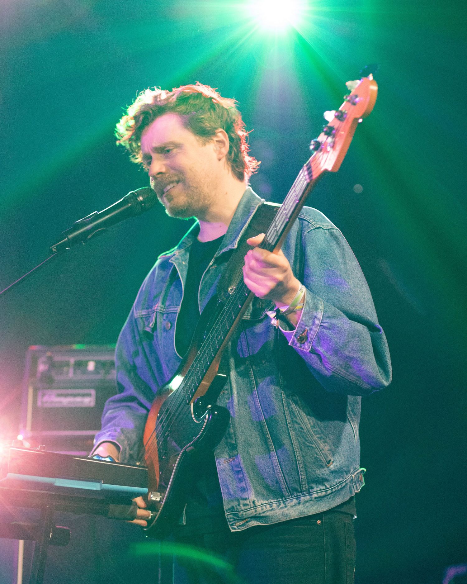 Male bassist smiling while playing electric bass guitar under green and purple stage lights.