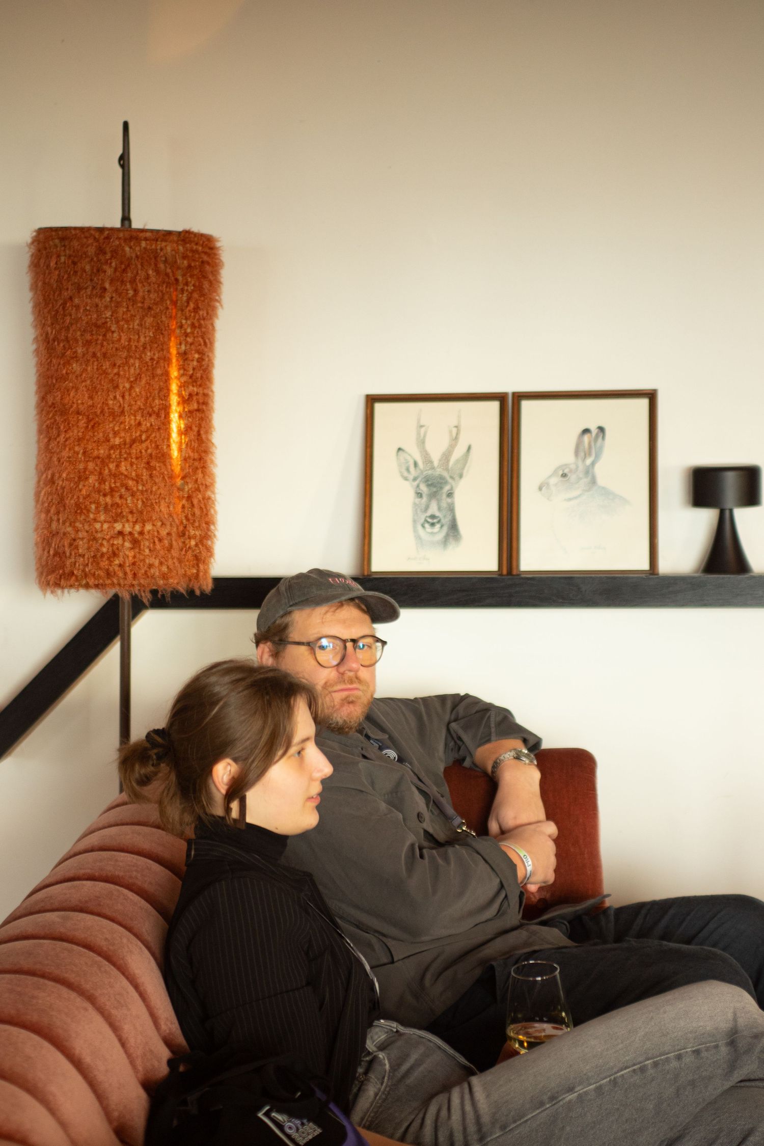 Couple relaxing on a pink sofa beneath framed deer and rabbit illustrations with an orange pendant lamp.