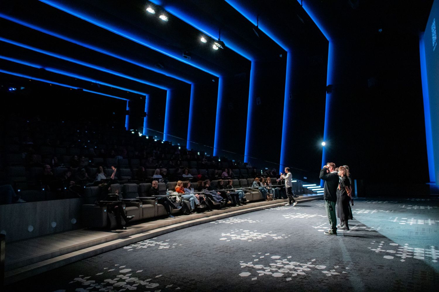 A spacious modern cinema auditorium with blue LED lighting frames two presenters standing on the stage floor.