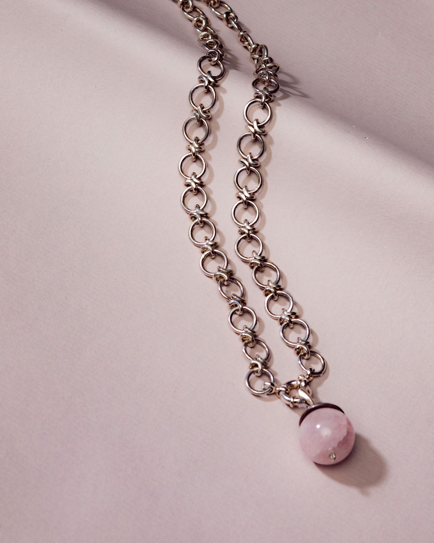 Silver chain necklace with a pink rose quartz ball pendant displayed on a light pink background.