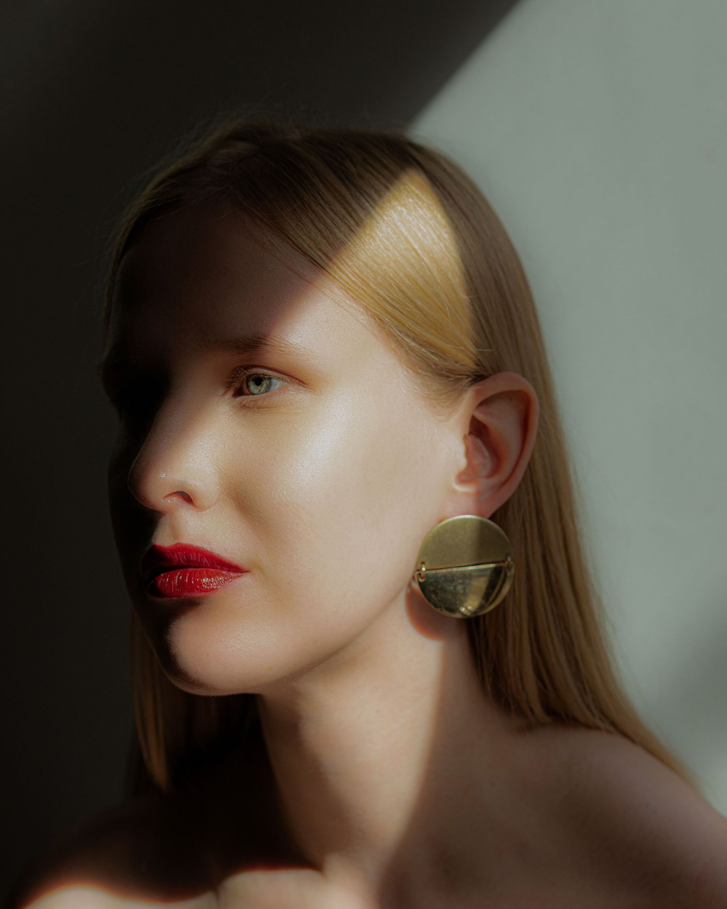 Blonde woman with red lips and large gold earring in a moody, shadow-lit portrait photo.