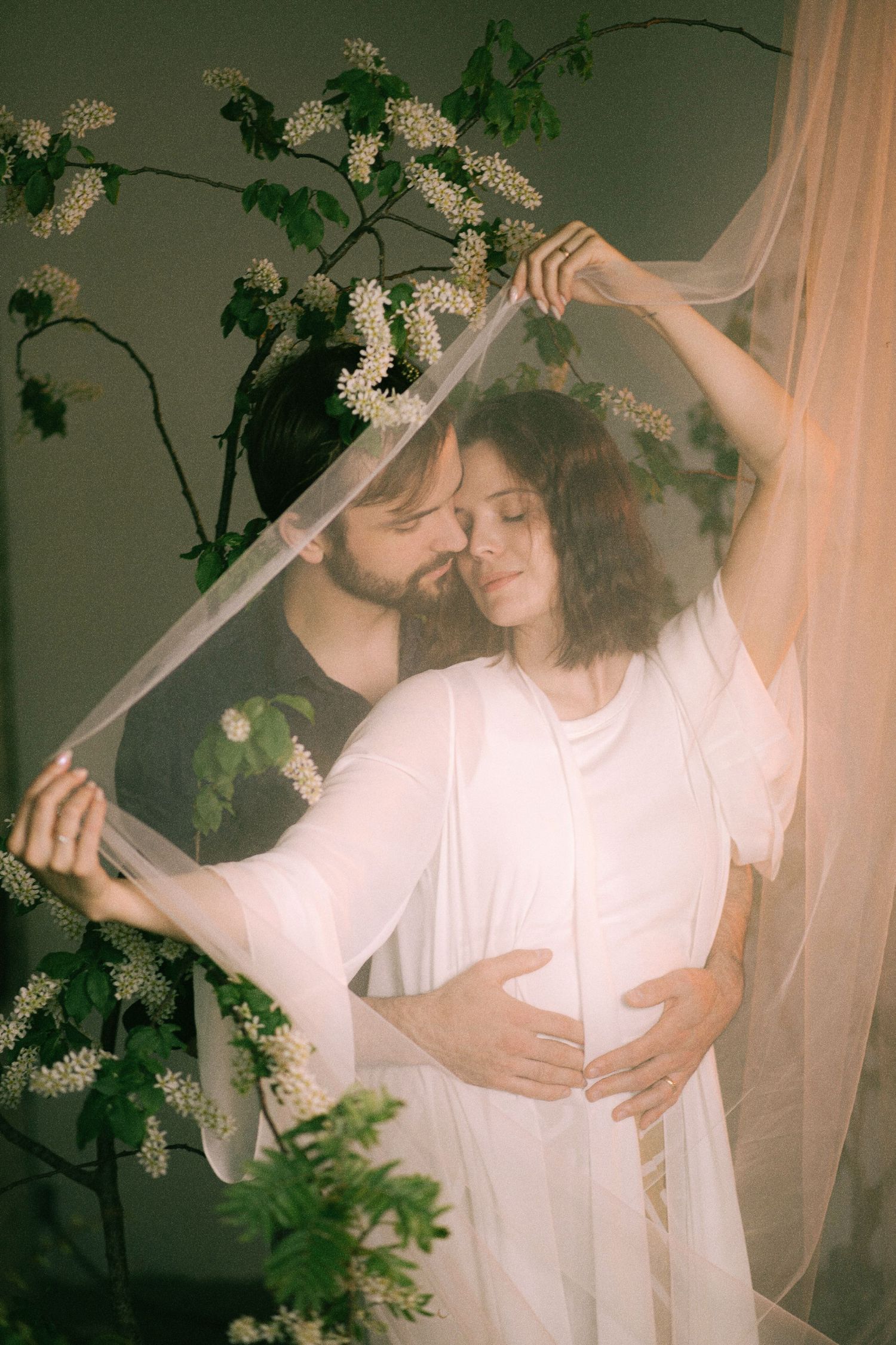 A couple embraces tenderly under a sheer veil surrounded by white blooming branches in a romantic maternity photo.