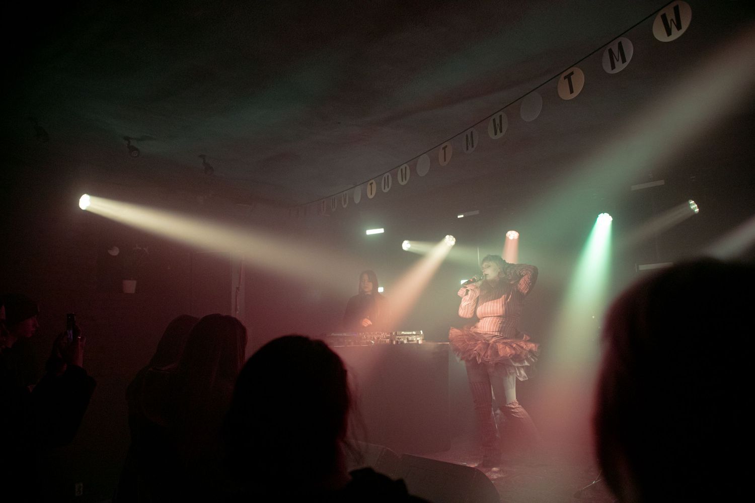 Live concert performance on stage with colorful spotlights illuminating performers before a crowd in a dimly lit venue.