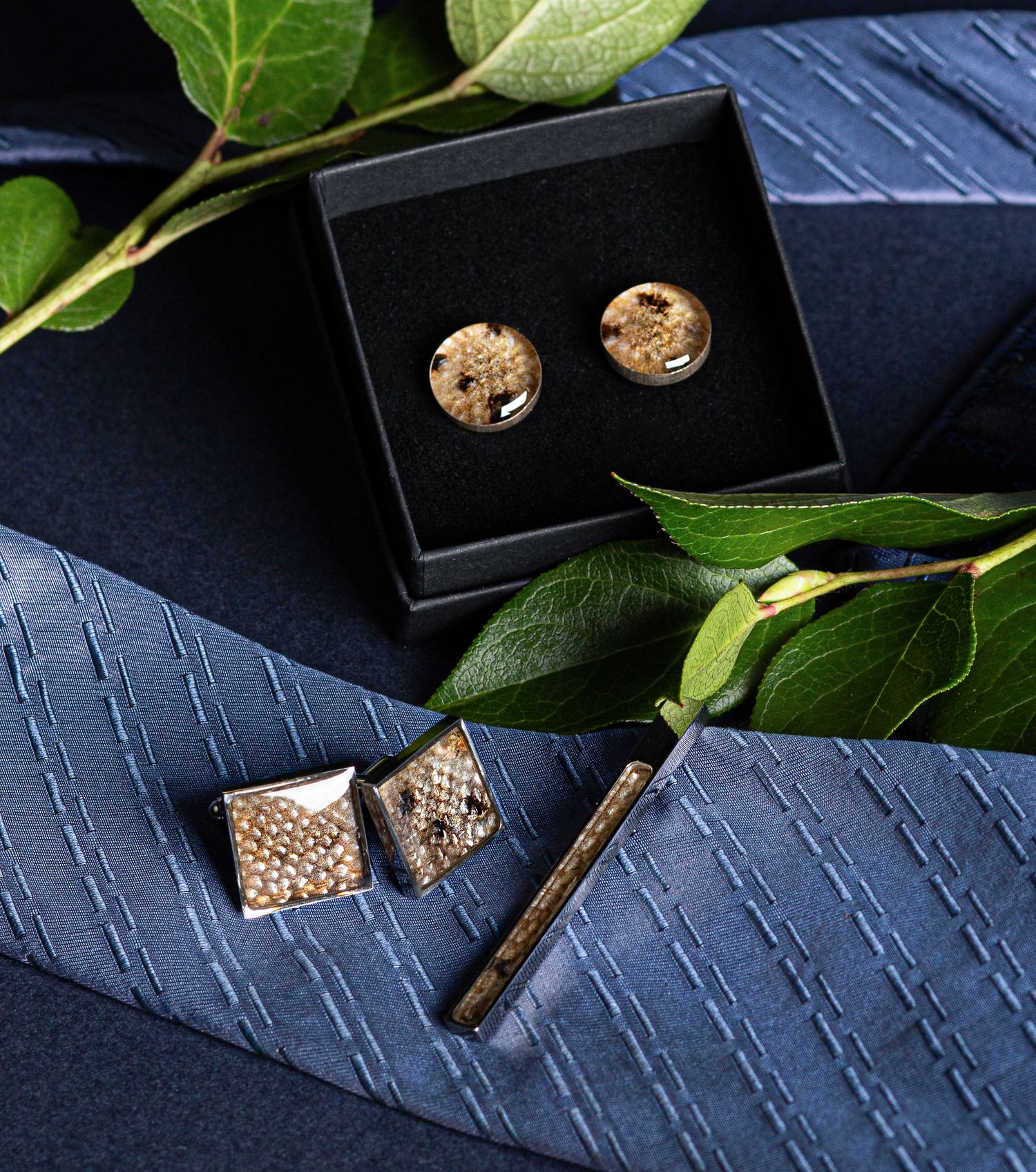 Gold cufflinks and tie bar set displayed on a navy blue silk tie with elegant gift box and green leaves.