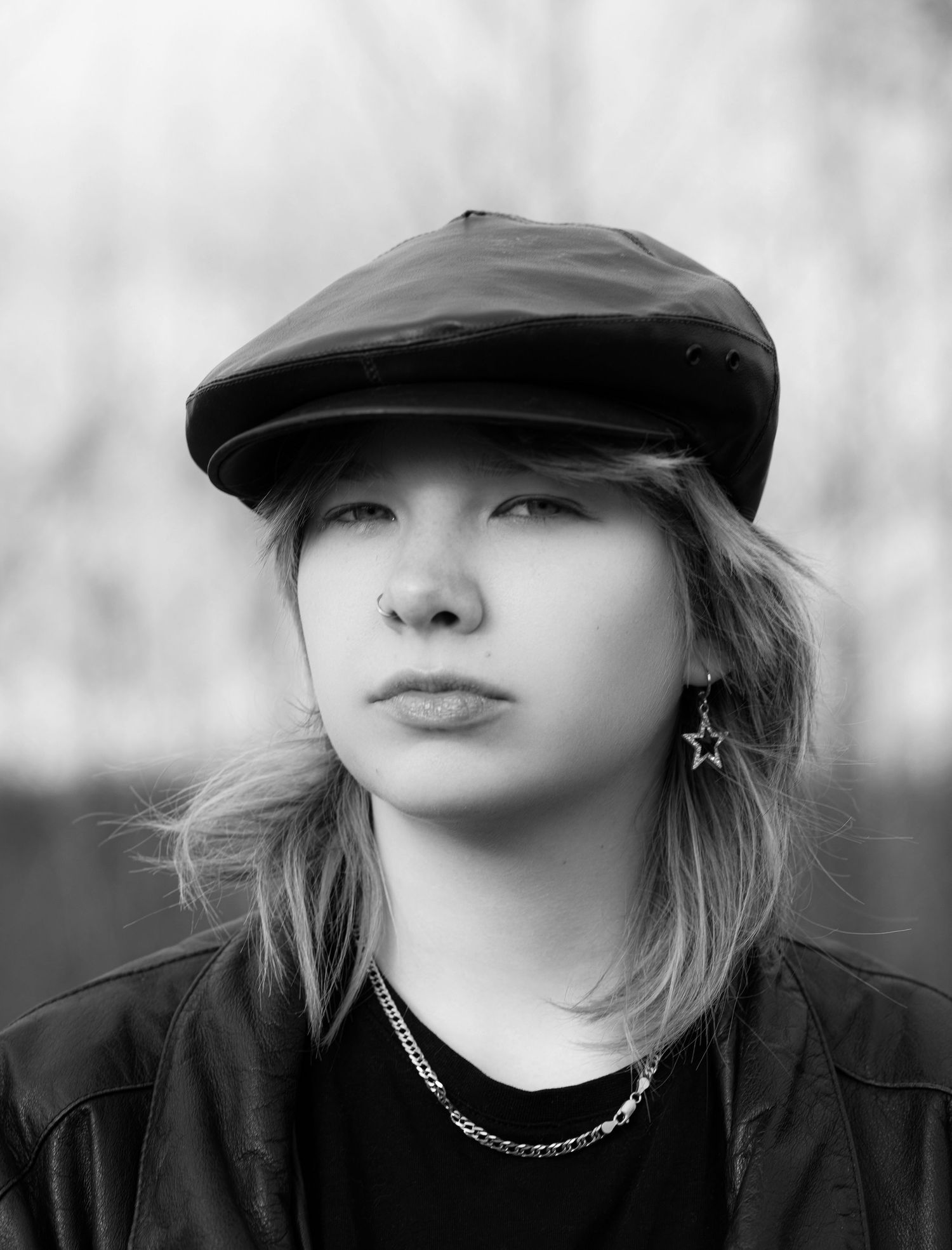 Black and white portrait of a young woman wearing a leather flat cap, star earring, and chain necklace.