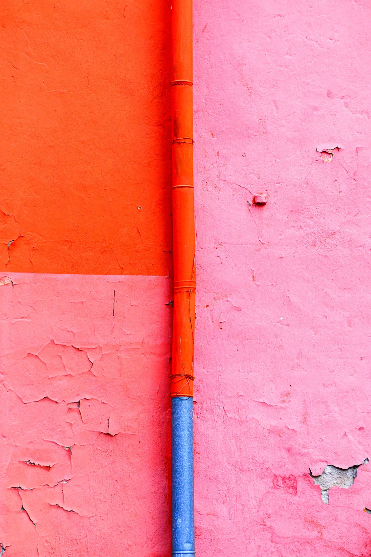 Vibrant orange and blue pipe running vertically along a textured pink and red painted wall.
