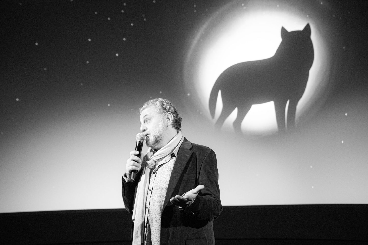 A black and white photo of a man speaking into a microphone before a projected wolf silhouette under a full moon.