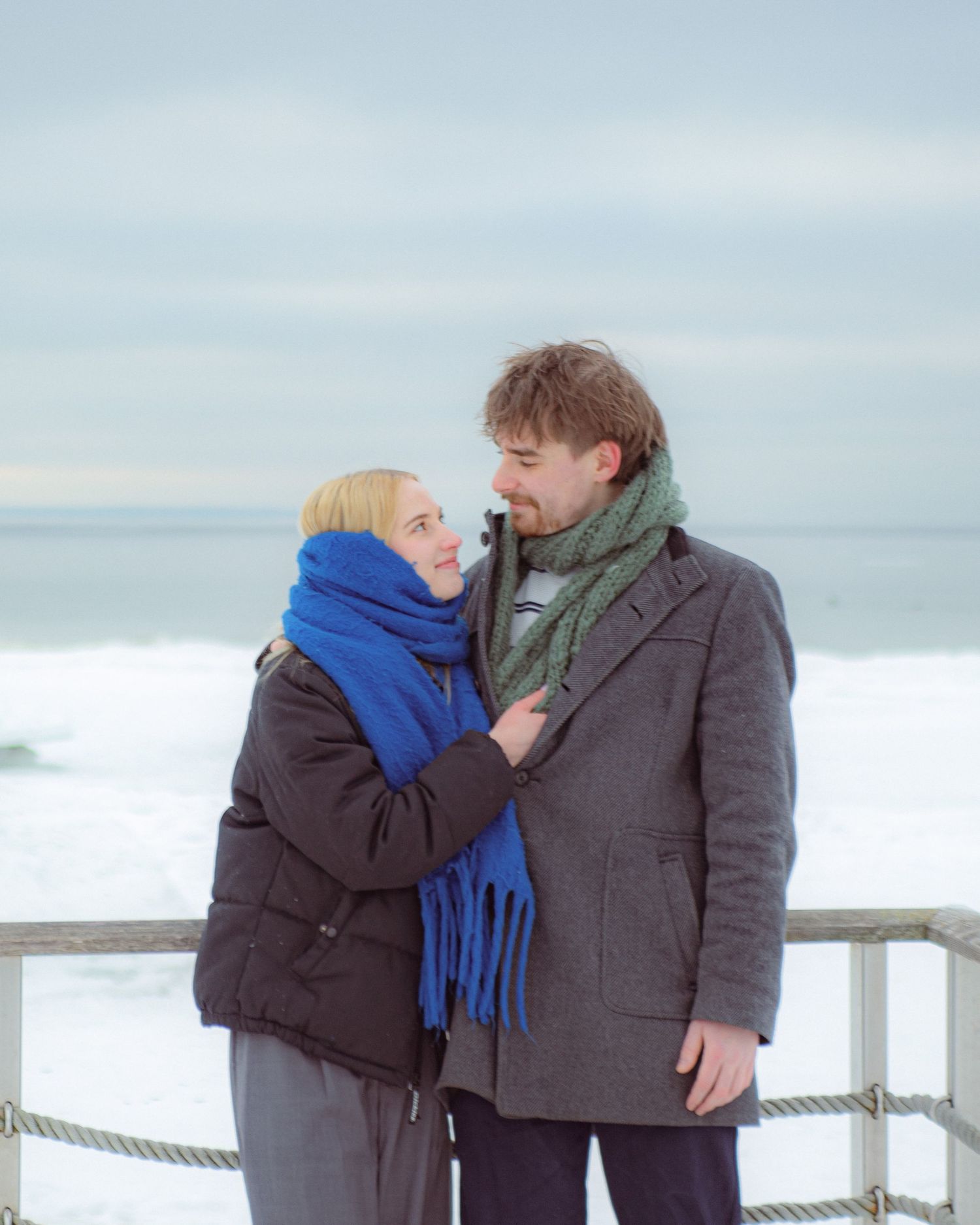 Couple in winter scarves smiling at each other on a snowy beachside boardwalk.