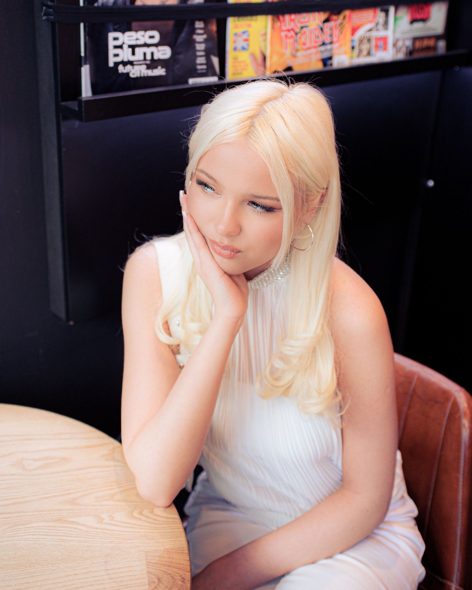 Young woman with long blonde hair wearing a white dress, resting her chin on her hand in a stylish indoor setting.