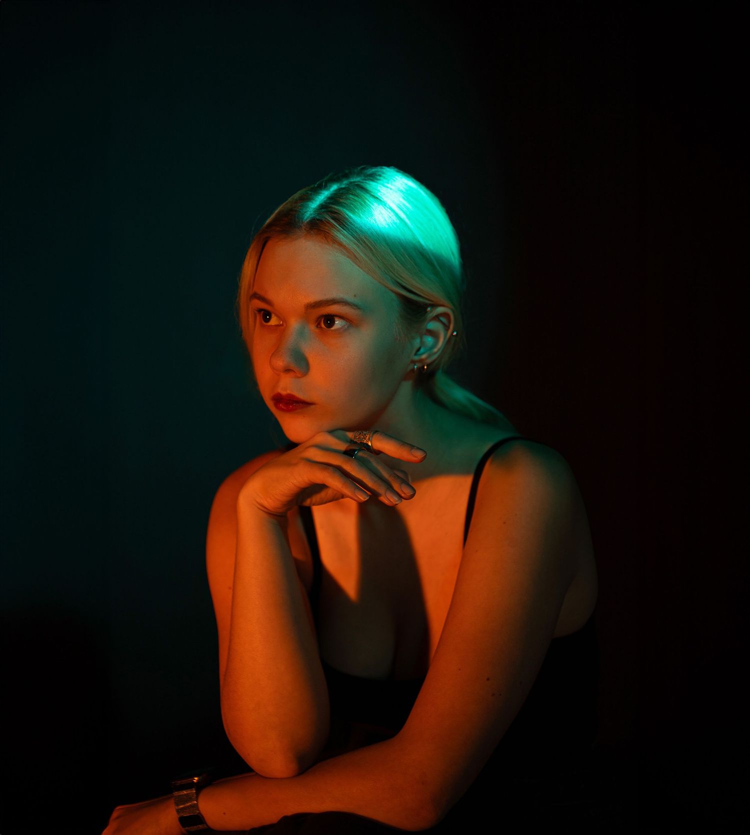 Young blonde woman poses thoughtfully under teal and orange neon lighting in dark studio portrait.