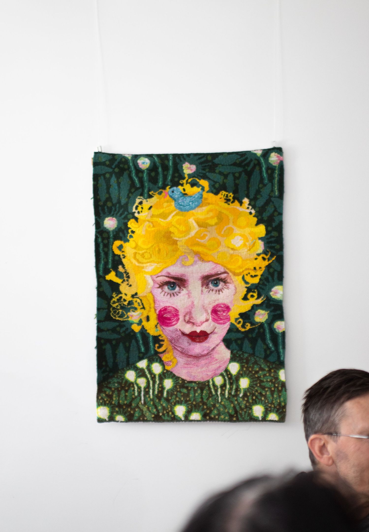 Colorful textile art portrait of woman with yellow curly hair and pink roses on white wall.