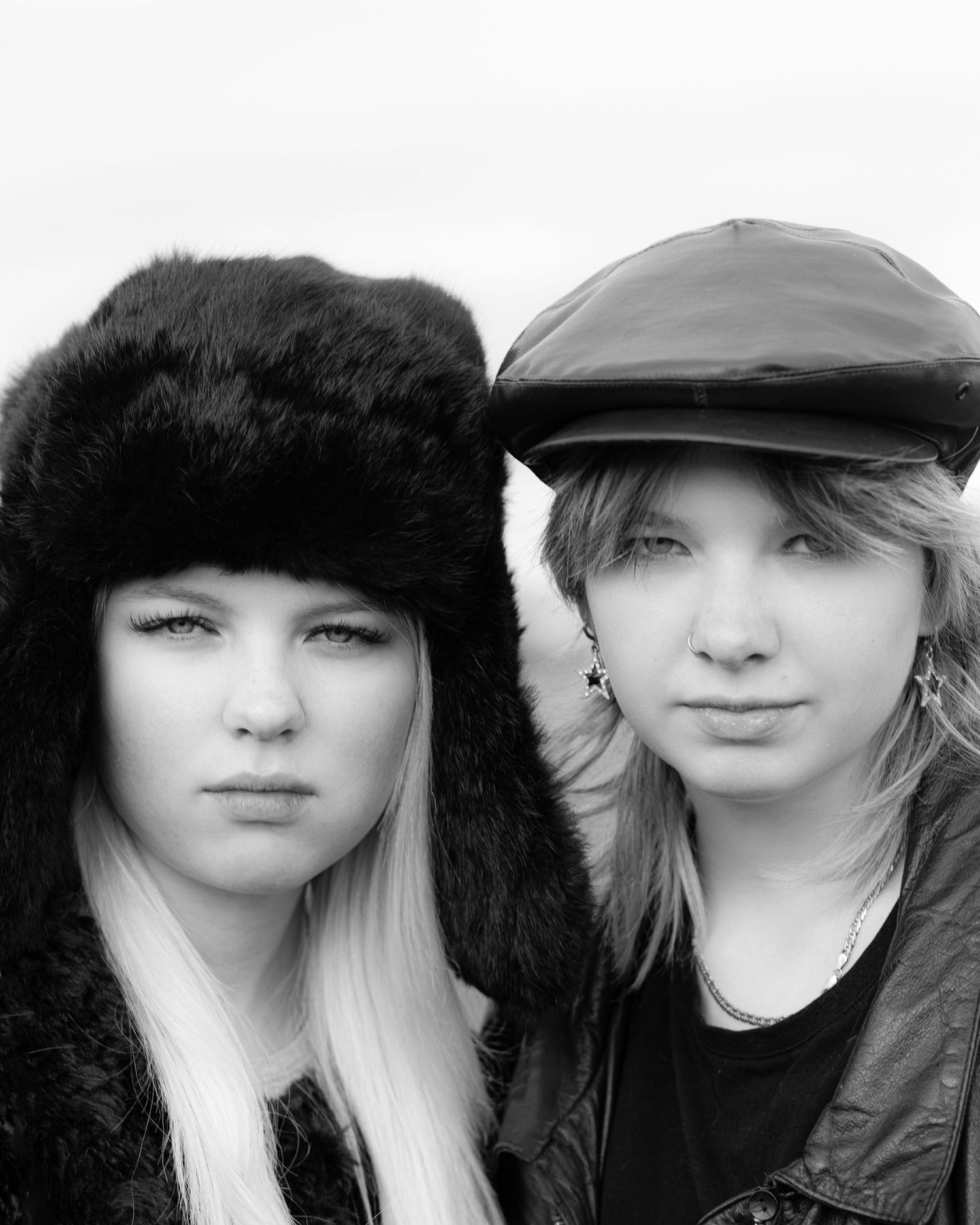 Black and white portrait of two young women wearing stylish hats, one fur and one leather cap, outdoors.