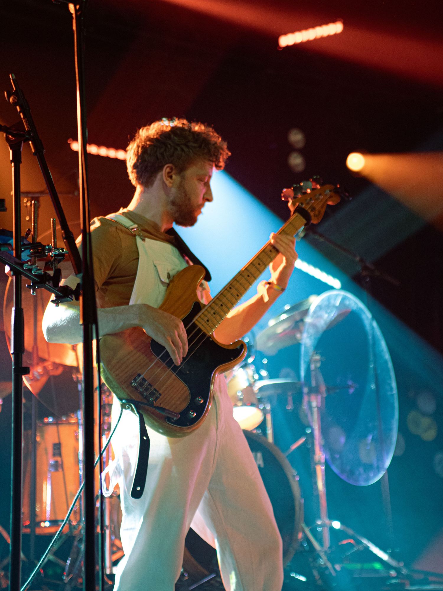 Male bassist playing electric guitar on a colorfully lit concert stage in white pants.