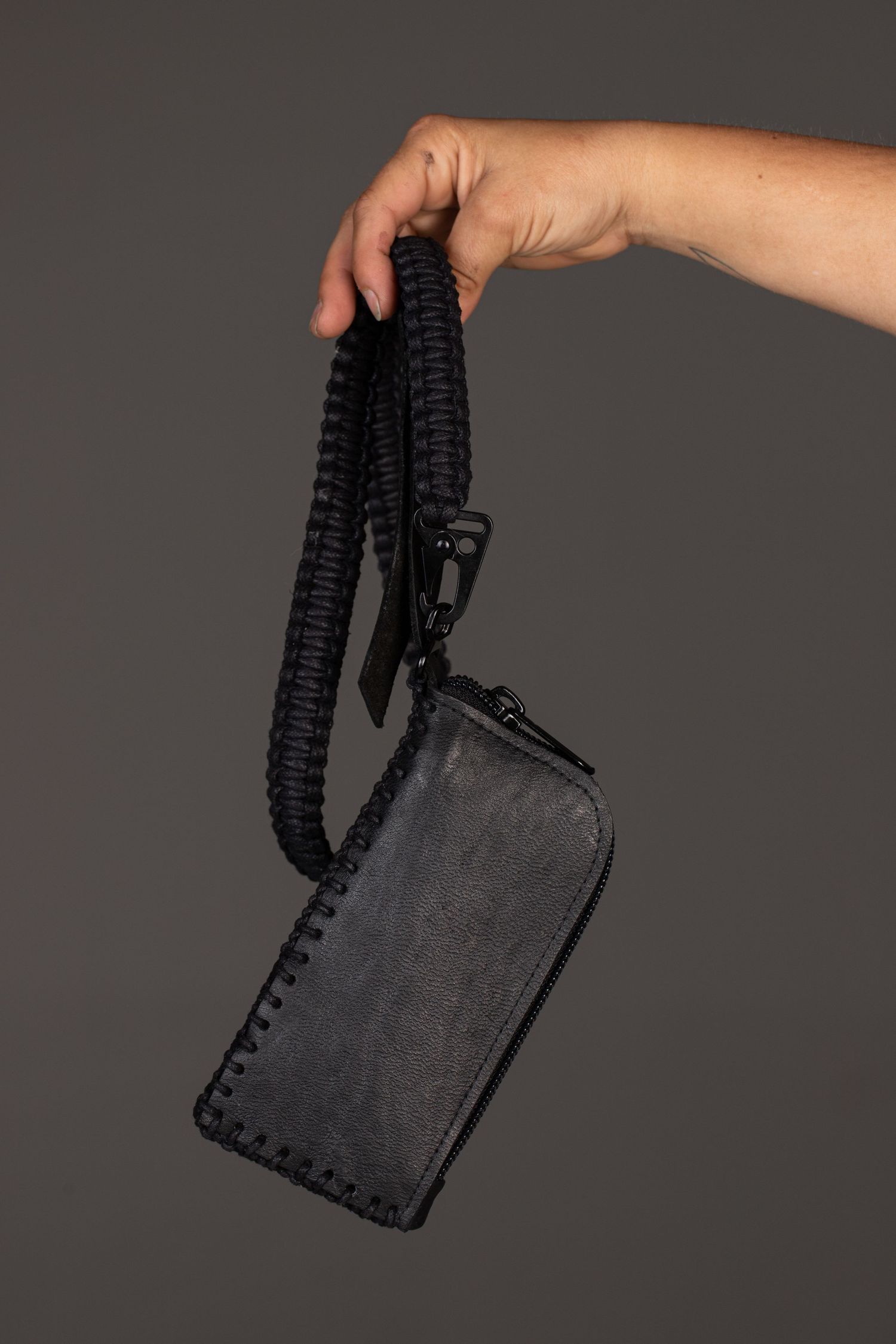 Hand holding a black leather wristlet wallet with braided paracord strap on gray background.
