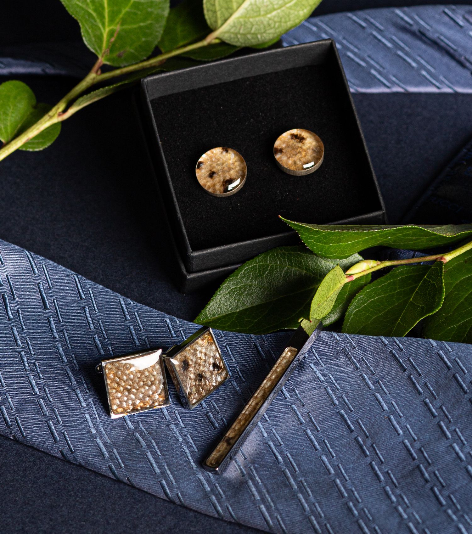 Elegant gold cufflinks and tie bar set displayed in a black jewelry box with green leaves and blue tie.