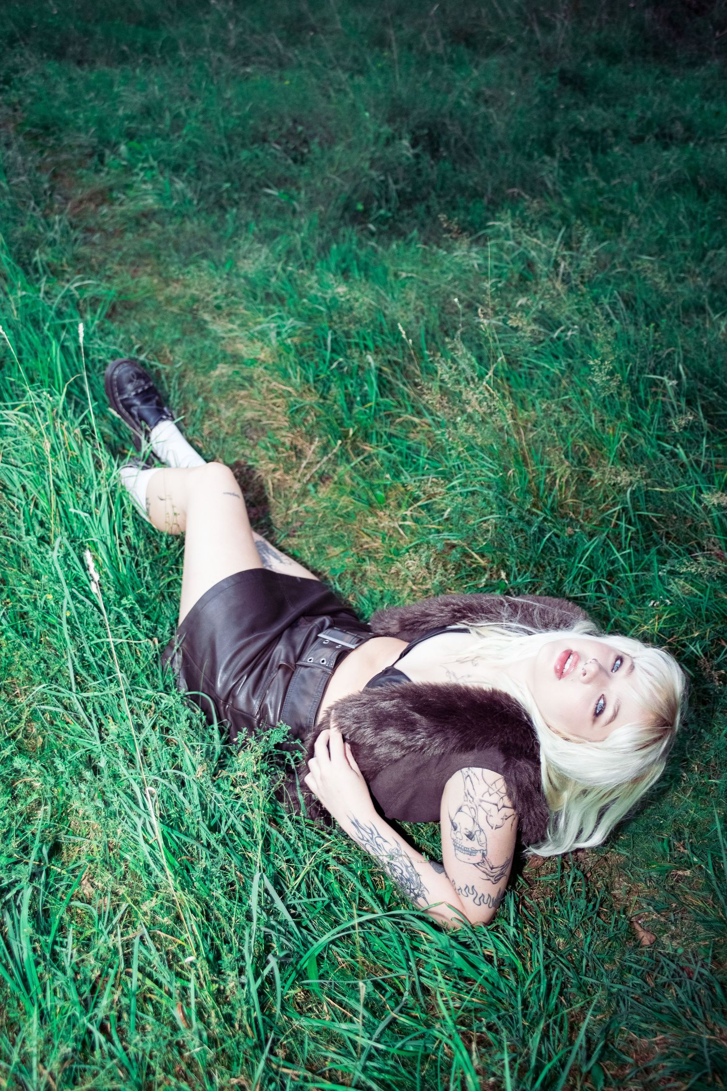 Platinum blonde woman with tattoos in a dark outfit lying in lush green grass, relaxing outdoors.
